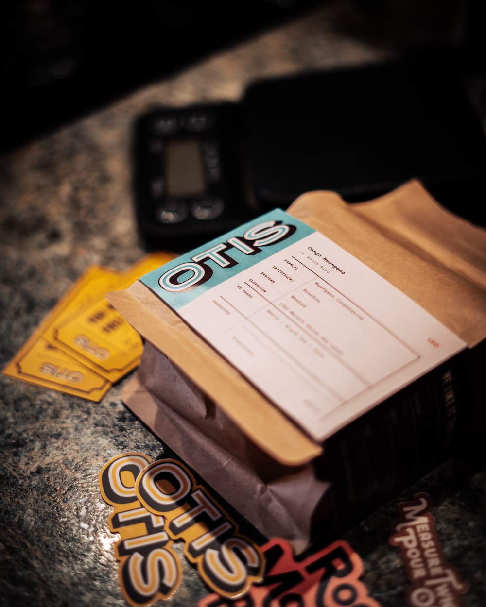 Single origin specialty coffee from OTIS with free drink cards and stickers on a counter next to a scale